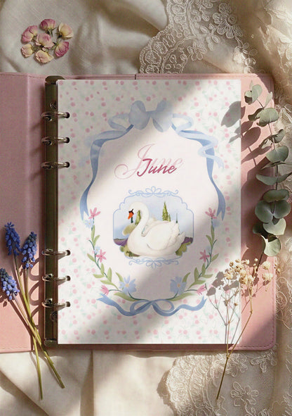 June Bullet Journal cover page inside a pink leather binder, featuring a graceful swan illustration framed by blue ribbons and delicate floral patterns. Top-down lifestyle photo with dried flowers, eucalyptus, and lace fabric in soft natural sunlight.